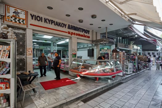 Crete, Greece - April 5, 2025: A bustling butcher shop with colorful displays of meat cuts, a red carpet entrance, and shoppers interacting near stalls and shops in a lively market.