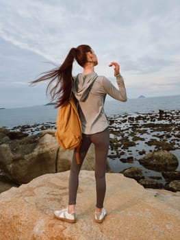 An athletic young woman with long hair, dressed in sportswear, stands on rocky shore, embracing nature and adventure with a backpack, showcasing a peaceful coastal scene