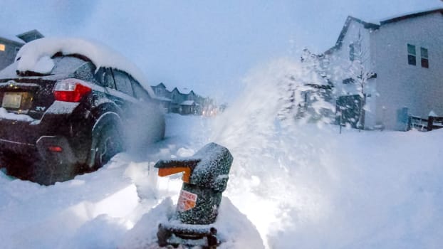 Denver, Colorado, USA-February 4, 2024-Snow blower is at work in the early evening light, clearing a path in a neighborhood where houses and a large snow pile are softly illuminated against the dusky sky.