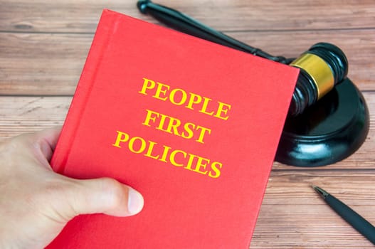 Hand holding People First Policies book with gavel on a wooden table background.