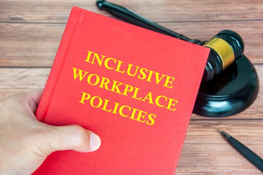 Hand holding Inclusive Workplace Policies book with gavel on a wooden table background.