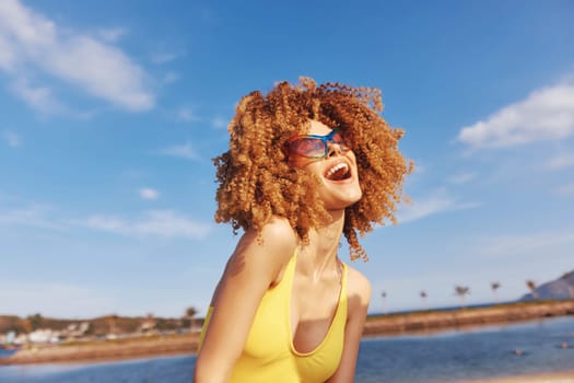 A cheerful young woman with curly hair, wearing sunglasses, smiles at the beach, with a clear blue sky and water in the background