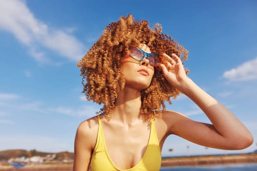 A vibrant scene of a cheerful woman with curly hair, dressed in a yellow swimsuit, relaxing on a sunny beach with clear blue skies and soft clouds