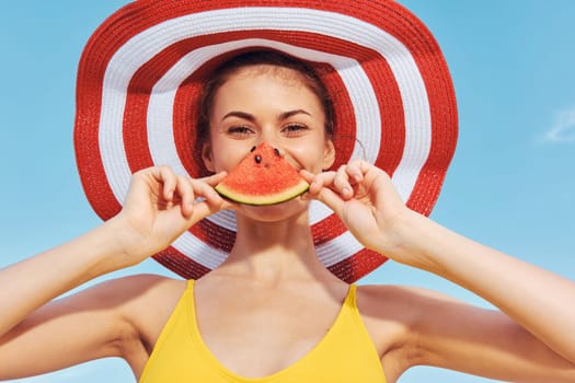 A joyful young woman in a yellow swimsuit poses under a big striped hat, playfully holding watermelon, enjoying summer vibes