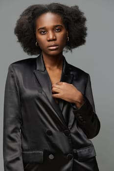 A confident young woman with natural afro hair poses in a stylish black blazer, set against a muted grey background, representing modern elegance and empowerment