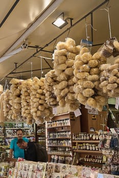 Crete, Greece - April 5, 2025: Natural sea sponges hanging in clusters at a bustling market stall, surrounded by honey jars, packaged olives, and signs indicating prices in a vibrant setting.