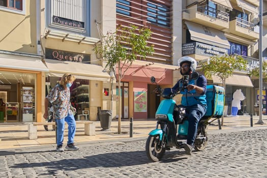 Crete, Greece - April 5, 2025: Dynamic image of a food delivery scooter with Wolt branding, surrounded by urban Mediterranean charm, yellow facades, and bustling street activity.