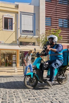 Crete, Greece - April 5, 2025: Vibrant moment of a delivery rider in uniform crossing cobblestones, carrying a recognizable Wolt bag, with classic European city elements in the background.