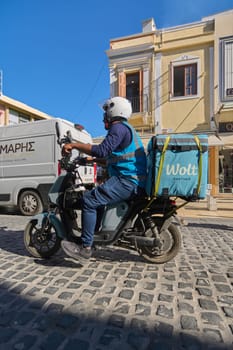 Crete, Greece - April 5, 2025: Wolt courier riding a scooter through a cobblestone street, carrying a branded delivery bag, against a backdrop of vibrant yellow buildings and clear blue skies.
