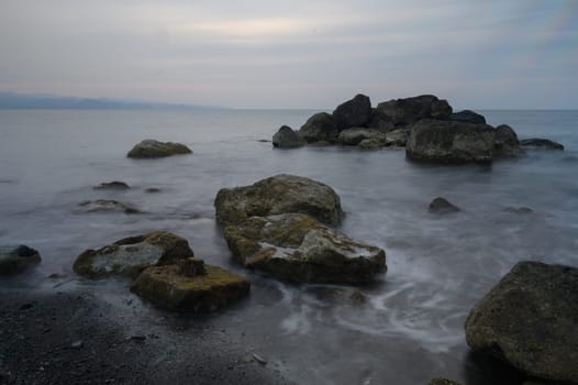 As dusk settles in, waves caress the rugged rocks along the shoreline, creating a soothing melody. The muted sky reflects a sense of calm over the serene coastal landscape.