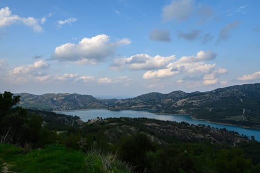 Bright clouds drift across a vivid blue sky above a tranquil lake surrounded by gentle hills. Nature unfolds in peaceful harmony under the warm sun, inviting relaxation and contemplation.