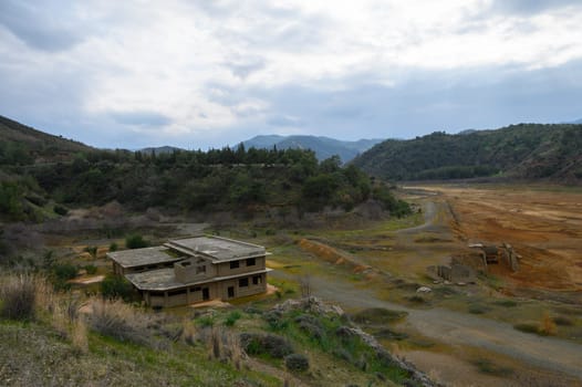 A derelict building stands isolated amidst a dry landscape, surrounded by rolling hills and a cloudy sky, evoking a sense of eerie abandonment and faded memories.