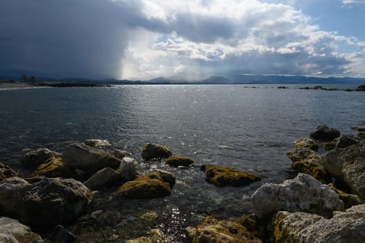 Waves lap gently against the moss-covered rocks as dark clouds gather over distant mountains during twilight. The reflective water shimmers under the changing light, creating a serene atmosphere.