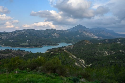 Bright clouds float above a serene lake surrounded by verdant hills and majestic mountains under a soft afternoon light, creating a peaceful atmosphere in nature.