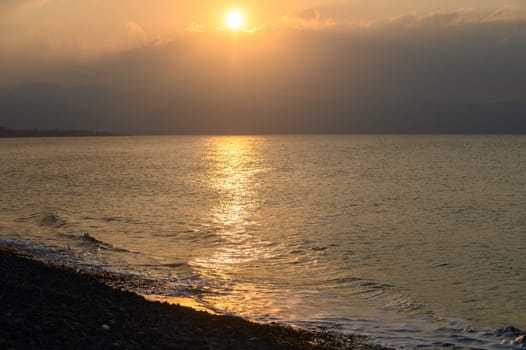 Waves gently lap against a pebbled shore as the sun sets on the horizon, creating a breathtaking reflection of gold and orange hues across the water. Nature's beauty unfolds in this peaceful moment.