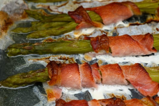 Fresh asparagus spears are delightfully wrapped in crisp bacon, sizzling and browning on a baking sheet. The warmth of the kitchen adds to the inviting aroma of this delicious dish.