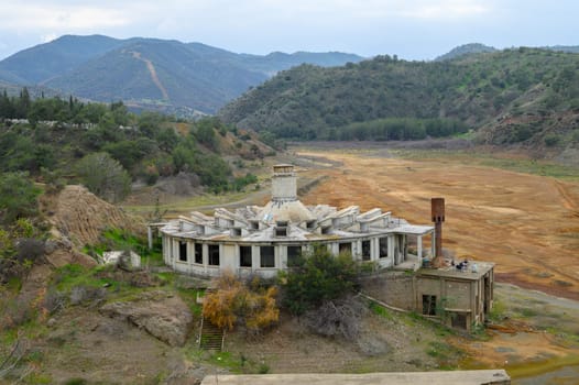 Decaying building stands alone by a barren riverbank, surrounded by rolling hills under a cloudy sky. Nature reclaims this forgotten site, evoking a sense of intrigue and nostalgia.