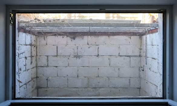 View through a window showing a small, dirty basement with white brick walls, a rusty metal grate, and spiderwebs, suggesting neglect and disrepair