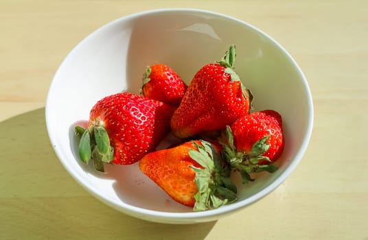 Five ripe strawberries with green leaves sit in a white bowl, illuminated by natural light on a wooden surface