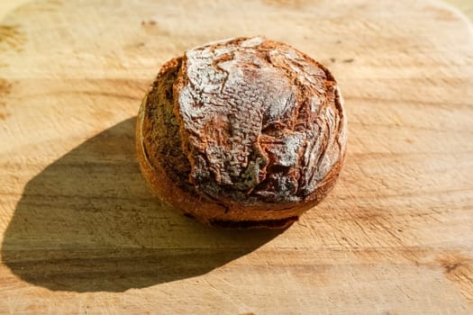 Freshly baked round sourdough bread loaf resting on a wooden cutting board, casting a strong shadow in natural light