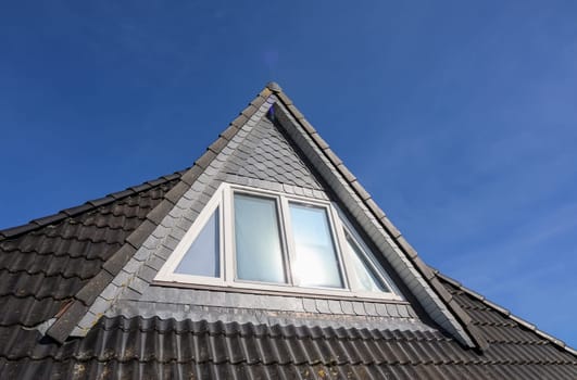 Triangular dormer window with white frame reflecting sunlight on a slate tiled roof of a residential building against a clear blue sky