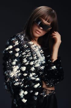 Woman wearing sunglasses and sequin jacket posing against a black background with her hand on her hip, exuding confidence and glamour in a stylish, highfashion shoot