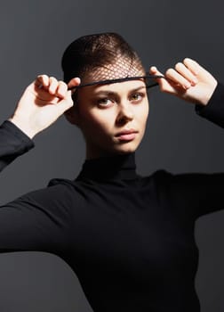 Confident female model dressed in a black turtleneck and fishnet headwear, expressing strength and individuality against a dark and simple background. Minimalist fashion and beauty concept.