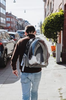 Knee length portrait of young caucasian stylish guy from the back who is walking on foot along the city street with a black and white cat in a transparent backpack on her back one spring sunny day, side view, close-up. People and cats concept, pet travel time, vet visit.