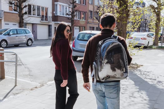 Portrait of young caucasian stylish happy couple having fun walking around the city with black and white cat in transparent backpack on their back one spring sunny day, side view, medium shot. Concept of people and cats, pet travel time, visit to vet.