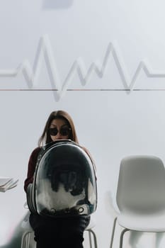 Portrait of one young beautiful Caucasian brunette girl in sunglasses sitting on a white chair in a veterinary clinic with a black and white cat in a transparent backpack on her lap waiting for an appointment spring day, side view, close-up. Concept of people and cats, visit to the veterinarian.