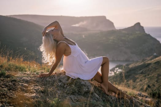 A woman is sitting on a hillside overlooking the ocean. She is wearing a white dress and has blonde hair. The scene is serene and peaceful, with the ocean in the background