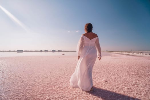 A woman in a white dress stands with her arms raised, facing away from the camera. She stands on a pink salt flat with a blue sky in the background.