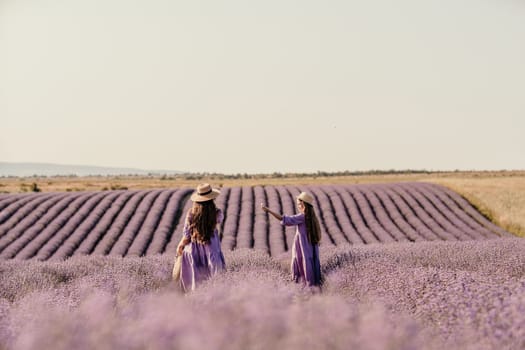 Mom and daughter are running through a lavender field dressed in purple dresses, long hair flowing and wearing hats. The field is full of purple flowers and the sky is clear