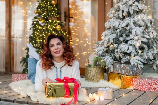 A woman with red hair smiles as she holds a wrapped present in front of a Christmas tree. She is sitting on a porch surrounded by presents and Christmas lights.