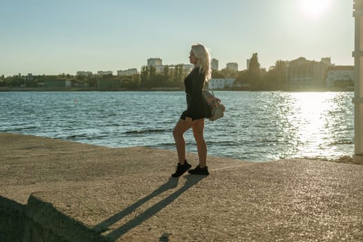 A woman in a black dress stands on a pier overlooking a body of water, with a city skyline in the background. The sun is setting, casting a warm glow on the scene.