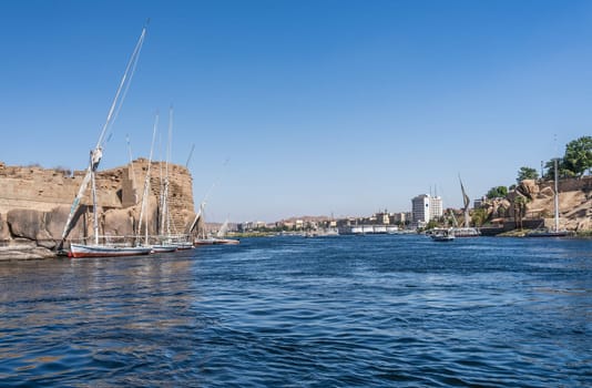 Skyline of Aswan in distance with Felucca boat docked by Ruins of Abu and many river cruise boats docked