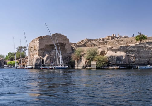 Felucca boat docked by Ruins of Abu outside Aswan in Egypt and other river cruise boats docked by the massive stone walls
