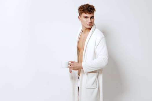 Confident young male in a cozy white robe enjoying a cup of coffee, showcasing a relaxed and trendy lifestyle in a minimalist indoor environment.