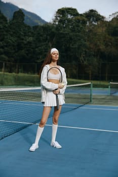 Confident and cheerful young woman dressed in trendy tennis apparel, standing on a vibrant blue court, embodying a passion for sports and health in a natural setting