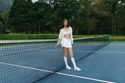 A confident young female tennis player dressed in white sportswear stands on a tennis court, exuding enthusiasm and joy The lush green background enhances the vibrant outdoor atmosphere, perfect for a summer sports concept