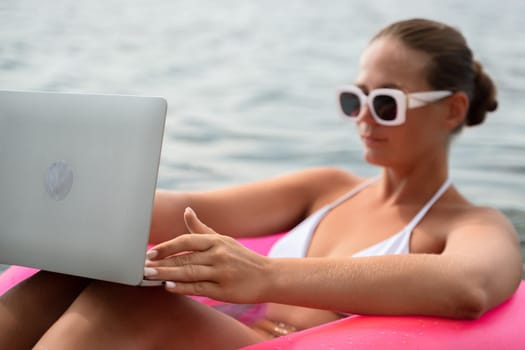 A woman is sitting in a pink inflatable raft on a lake, holding a laptop. Concept of relaxation and leisure, as the woman enjoys her time in the water while working on her laptop