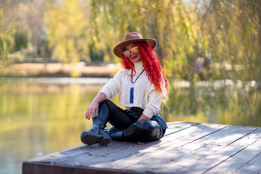Autumn lake woman. In autumn, she sits by the pond on a wooden pier and admires nature with red hair and a hat. Tourism concept, weekend outside the city