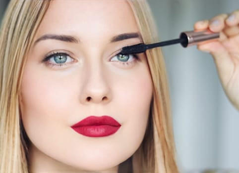 Woman applying mascara with confidence, defined brows and red lipstick. Blonde beauty focused on her eye makeup, cosmetics look