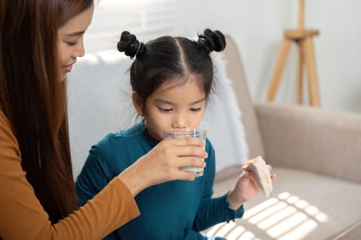 In a cozy home setting, a mother helps her daughter drink milk as she enjoys her breakfast, encapsulating the affection and care in their morning routine before school.