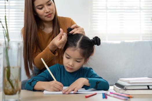A nurturing parent styles their daughter's hair while she concentrates on her homework, highlighting the balance between care and education in daily life.