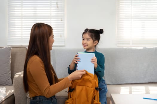 The little girl is excitedly holding her school supplies as her mother prepares her backpack, showcasing their school readiness.