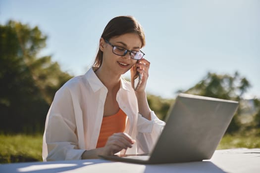 A young woman wearing glasses is sitting outdoors, engaged in a phone conversation while working on her laptop Her casual outfit includes a white shirt and orange top, highlighting a cheerful and pr