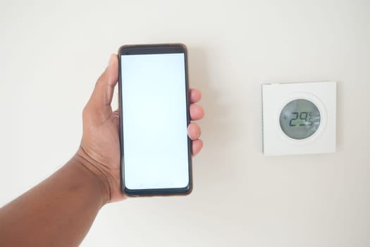 A person holds a smartphone by a digital thermostat showing temperature.
