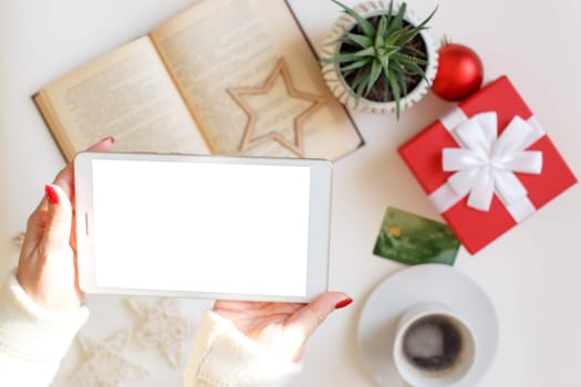 Beautiful woman hands hold digital tablet over table with credit card, open book, red gift box, cup of coffee and succulent plant, christmas decor and toys. Mock up