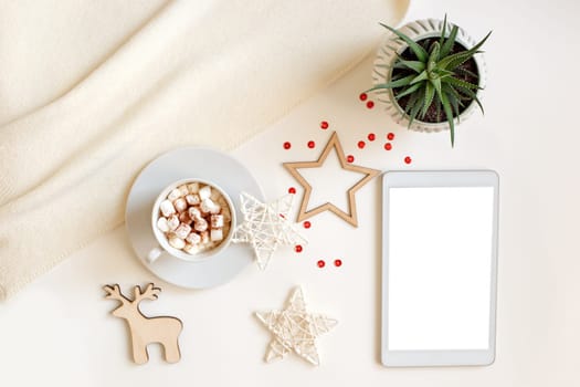 A digital tablet with a blank screen lies on a white table surrounded by flat wooden Christmas decorations and a white cup of coffee or cocoa with marshmallows, deer, stars. View from above.Mock up